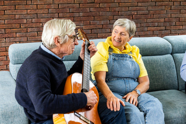 Radosny obraz przedstawiający starszego mężczyznę grającego na gitarze, podczas gdy kobieta, ubrana w żółtą koszulę i kombinezon, słucha z uznaniem. Para siedzi na wygodnej sofie na tle - Zdjęcie, obraz