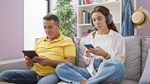 En casa, un padre cariñoso y su hija se relajan en un cómodo sofá, completamente absortos usando un touchpad y un teléfono inteligente, vinculándose por intereses compartidos en la tecnología. - Foto, imagen