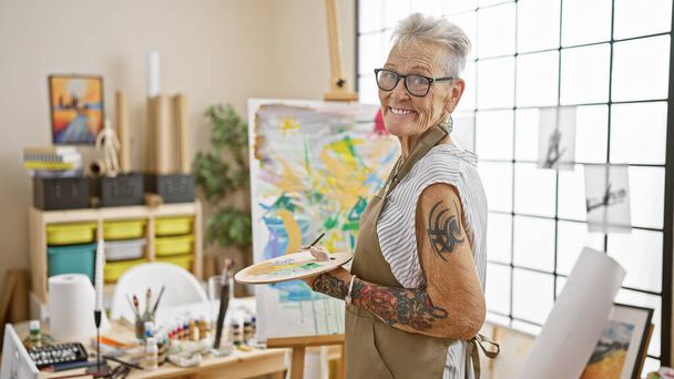Впевнена, сіро-волохата старша жінка художник, радісно посміхається, тримаючи її надійну пензлик і палітру в галасливій художній студії. - Фото, зображення