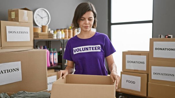 Poważna młoda Latynoska radośnie wolontariuszka w ośrodku charytatywnym, stojąca w pomieszczeniu z podarowanym pakietem, oferująca wsparcie społeczne z pięknym uśmiechem i krótką fryzurą - Zdjęcie, obraz