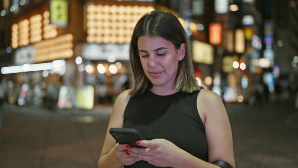 Mooie Spaanse vrouw verdiept in een serieus telefoongesprek, met behulp van haar smartphone op tokyo 's levendige straten onder de nachtlampjes van de stad - Foto, afbeelding