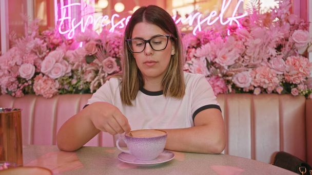 Young adult woman stirs coffee in a pink floral cafe, embodying a cozy, stylish ambiance. - Photo, Image