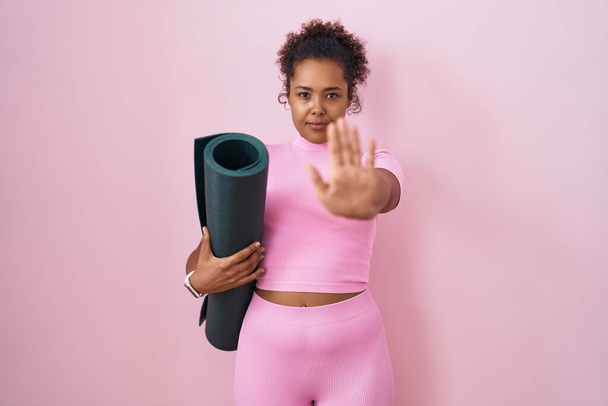 Jeune femme hispanique avec les cheveux bouclés tenant tapis de yoga sur fond rose faire arrêter de chanter avec la paume de la main. expression d'avertissement avec geste négatif et sérieux sur le visage.  - Photo, image
