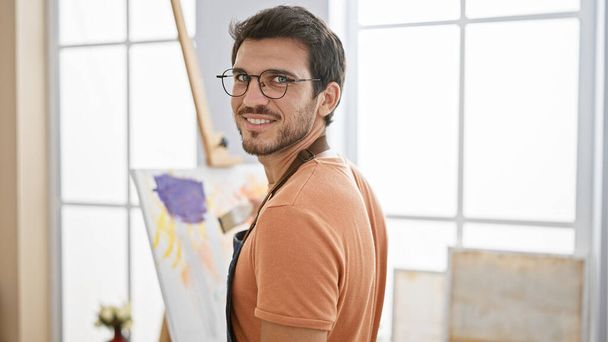 Egy mosolygó, szakállas spanyol fiatalember, szemüveget visel egy művészeti stúdióban, portréhoz pózol.. - Fotó, kép
