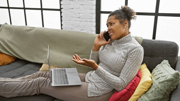 Una mujer afroamericana adulta expresa confusión mientras habla por teléfono, sentada en un sofá con una computadora portátil en el interior de un hogar moderno. - Foto, imagen