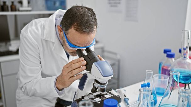 Зрілий чоловік з сірим волоссям- вчений, захоплений на роботі в лабораторії, заглиблений в медичні дослідження з використанням передових технологій мікроскопа. - Фото, зображення