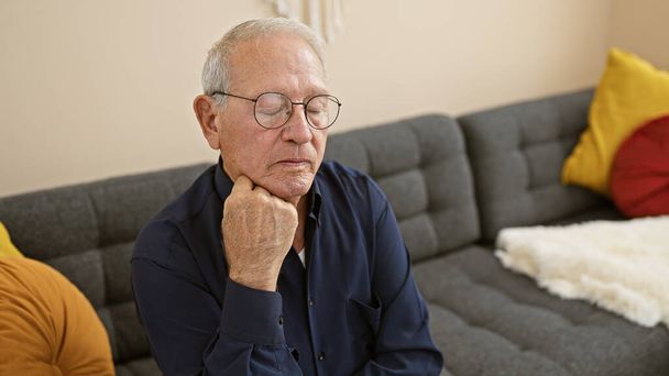 Preoccupato uomo anziano con i capelli bianchi al coperto, seduto sul divano del soggiorno a casa, mostrando un'espressione seria, piena di dubbio, sconvolto e tristezza, profondamente assorto nei suoi pensieri - Foto, immagini