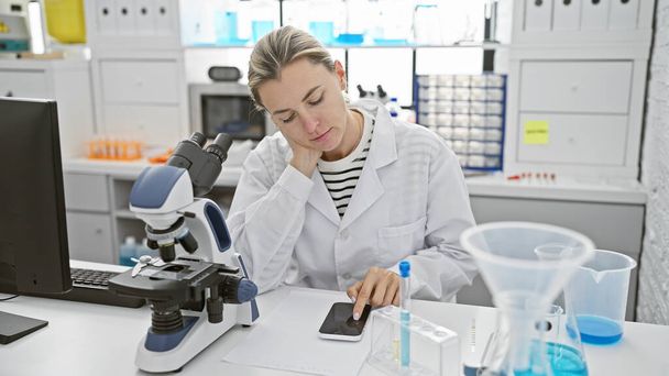 Una scienziata concentrata in un laboratorio utilizzando uno smartphone mentre lavora accanto a un microscopio e provette. - Foto, immagini
