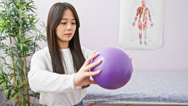 Молода азіатка тримає фіолетовий тренувальний м'яч у клініці з анатомічними графіками на задньому плані. - Фото, зображення