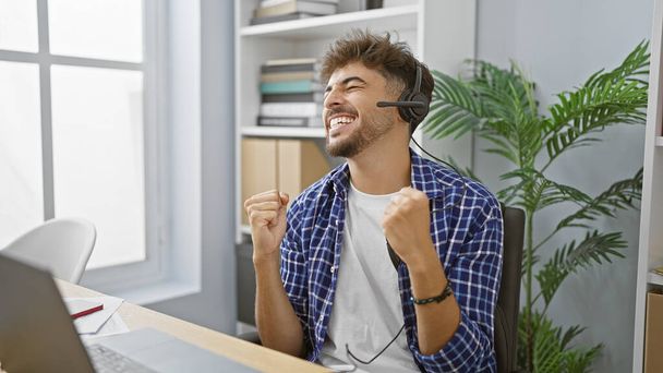 Ganar en el trabajo, joven árabe, un trabajador profesional, radiando positividad mientras celebra su éxito en la oficina, portátil en el escritorio y auriculares en - Foto, Imagen