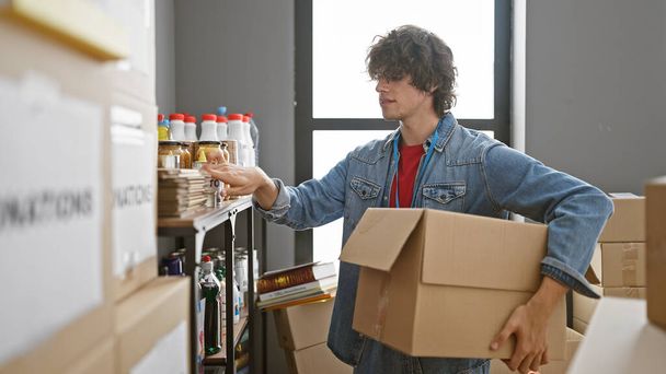 Un jeune homme aux cheveux bouclés, vêtu d'une veste en denim, emballe des produits dans un entrepôt avec des étagères en arrière-plan. - Photo, image