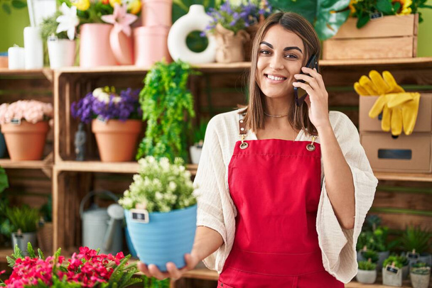Молода красива іспаномовна жінка-флорист розмовляє на смартфоні тримає завод в квітковому магазині
 - Фото, зображення