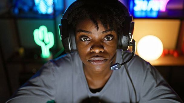 Una joven negra enfocada con un auricular se sienta en una sala de juegos iluminada por neón, ojos amplios con concentración. - Foto, Imagen