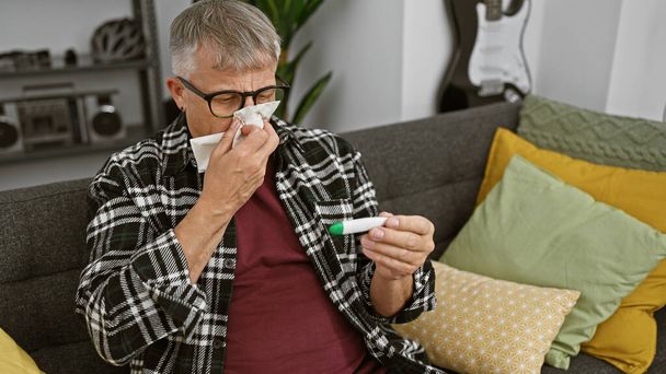 Um homem maduro com cabelos grisalhos senta-se em um sofá em casa, assoando o nariz enquanto verifica um termômetro, indicando doença. - Foto, Imagem