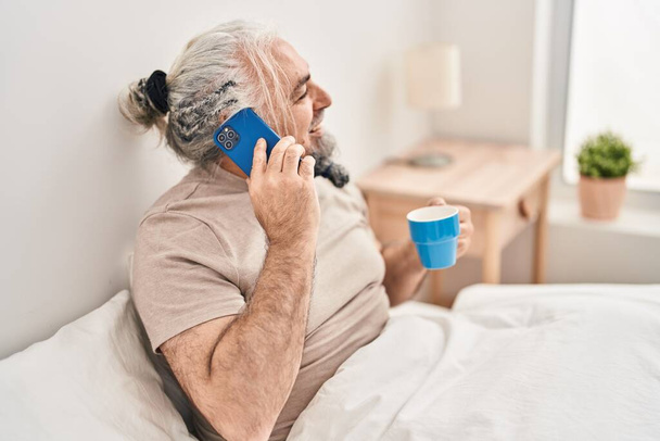 Седовласый мужчина средних лет разговаривает по смартфону и пьет кофе в спальне. - Фото, изображение