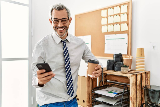 Trabajador de negocios hombre de pelo gris de mediana edad usando teléfono inteligente beber café en la oficina - Foto, Imagen