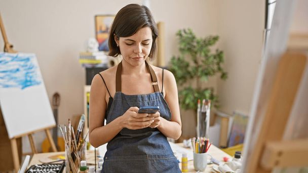 Young, beautiful, hispanic woman artist stands engrossed, smartphone in hand amid art studio canvas, paintbrush and palette, embracing the digital age of painting. - Photo, Image