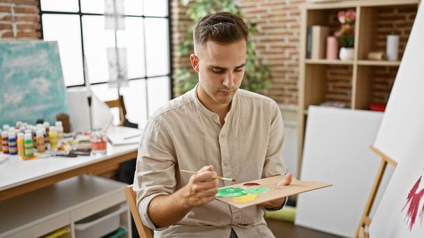 Un giovane uomo ispanico si concentra mentre dipinge su una tela in un ambiente di studio d'arte, irradiando vibrazioni creative e accademiche.. - Foto, immagini