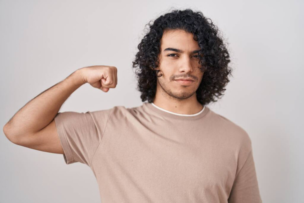 Homem hispânico com cabelos encaracolados em pé sobre fundo branco pessoa forte mostrando músculo do braço, confiante e orgulhoso do poder  - Foto, Imagem