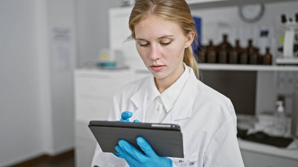 Vaalea nainen labratakissa keskittyy intensiivisesti tablettiin tieteellisen laboratorion sisällä, laitteiden ympäröimänä.. - Valokuva, kuva