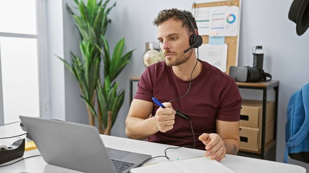 Egy spanyolajkú férfi fülhallgatót visel, egy irodában dolgozik laptoppal és jegyzetfüzetekkel.. - Fotó, kép