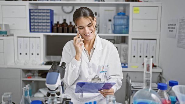 Stunning young hispanic woman scientist, reading report and talking on smartphone at lab work table, mastering the art of chemistry research - Photo, Image