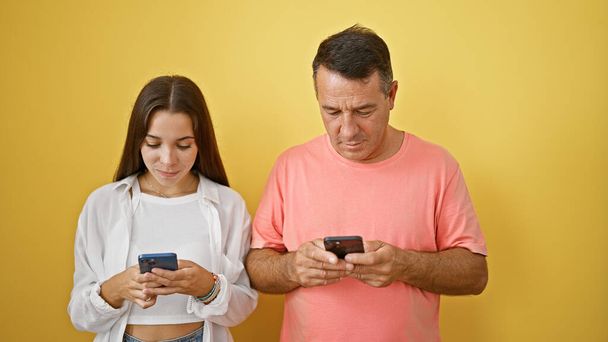 Isä ja tytär uppoutuivat älypuhelimiinsa eloisalla keltaisella, eristetyllä taustalla, kirjoittivat viestejä, navigoivat online-maailmassa, syvensivät yhteyksiään digitaalisen viestinnän avulla. - Valokuva, kuva