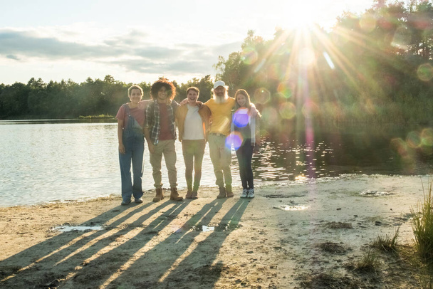 This photo captures a warm moment among a group of friends enjoying the golden hour by a serene lake. The sun, setting in the background, creates a radiant backdrop and casts long shadows on the sand - Photo, Image