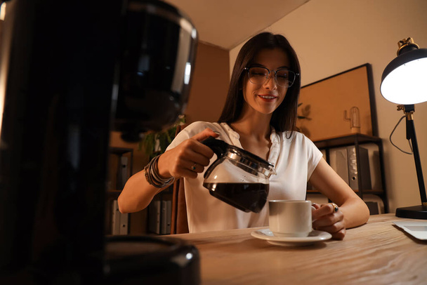 Mladá podnikatelka nalévání horké kávy do šálku v kanceláři v noci - Fotografie, Obrázek