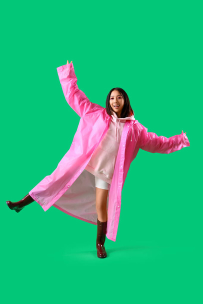 Young Asian woman in pink hoodie and raincoat on green background - Zdjęcie, obraz