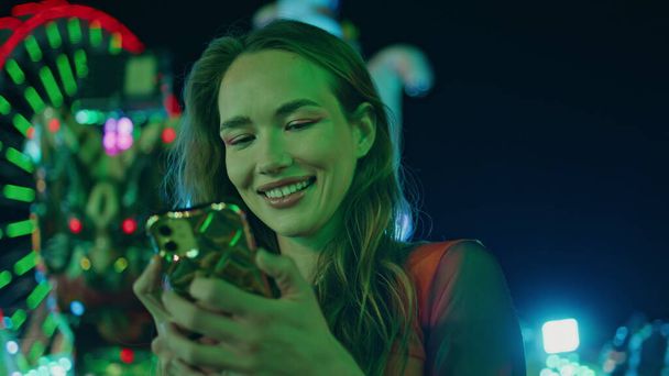 Νεόν κορίτσι μηνυμάτων smartphone closeup. Χαμογελώντας ελκυστική έφηβος γραπτών μηνυμάτων σε απευθείας σύνδεση φίλος αναμονή στο Λούνα Παρκ. Ευτυχισμένη γυναίκα που κρατάει το κινητό τηλέφωνο συζητώντας σε εφαρμογή ραντεβού. Όμορφο μοντέλο ανάπαυσης πόλη καρναβάλι - Φωτογραφία, εικόνα
