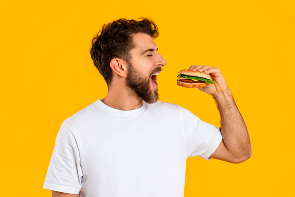 Портрет голодного парня, наслаждающегося сочным гамбургером на жёлтом фоне, молодого бородатого, наслаждающегося нездоровой едой, поедающего большой бургер. Концепция питания и нездорового питания - Фото, изображение