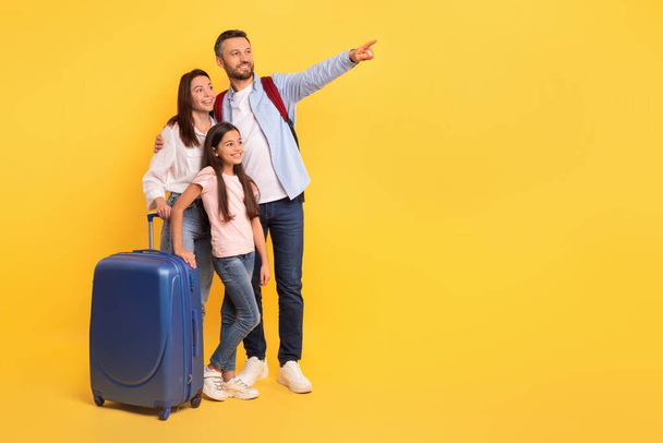 Family of three with suitcase ready for vacation, happily pointing to space for your travel offers against yellow studio background. Happy dad, mom and daughter with luggage showing copy space - Photo, Image