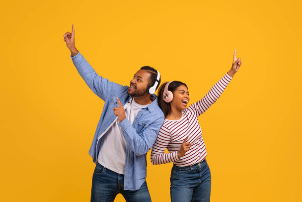 Šťastný mladý černošky pár nosí sluchátka těší hudbu a tanec spolu, veselý africký Američan muž a žena baví, stojí izolované proti pulzující žluté pozadí, kopírovat prostor - Fotografie, Obrázek