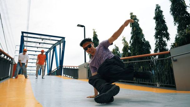 Panorama-Aufnahme von Hipster in stylischem Tuch und frierender Pose. Eine Gruppe professioneller Tänzer führt Breakdance auf, während der herausragende Mann B-Boy Dance übt. Outdoor-Sport 2024. Schwungvoll - Foto, Bild