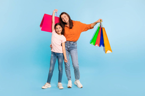 Onnellinen nuori äiti ja tytär poseeraa värikkäiden ostoskassien kanssa, nauttien onnistuneesta ostospäivästä, kantaen ostoja sinisellä taustalla, täyspitkä. Kausimyynti - Valokuva, kuva
