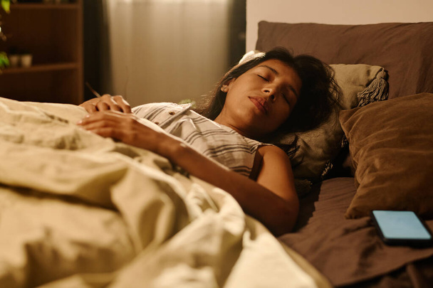 Νέοι κουρασμένοι Latino γυναίκα κρατώντας το κεφάλι σε μαλακό γκρι μαξιλάρι, ενώ βρίσκεται κάτω από κουβέρτα σε άνετο κρεβάτι και ύπνο τη νύχτα - Φωτογραφία, εικόνα