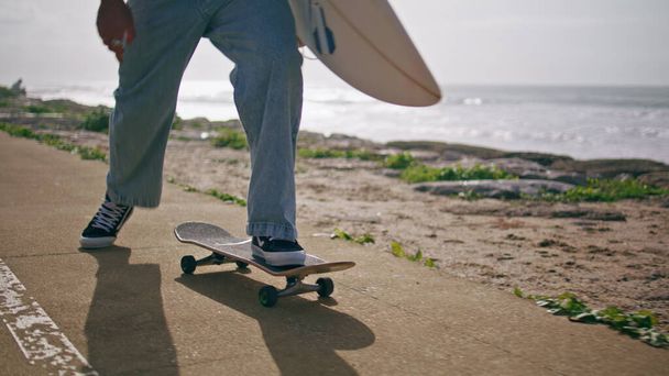 Unknown skater practicing skateboarding on road near beautiful sunny ocean view close up. Skateboarder legs pushing off asphalt accelerating on skateboard. Guy skating holding surfboard in slow motion - Photo, Image