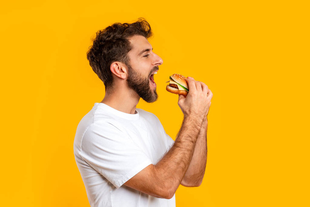 Портрет венгра в белой футболке, кусающегося в студии на желтом фоне, поедающего вкусный чизбургер в качестве закуски. Junk Food For Comfort, несбалансированное питание - Фото, изображение