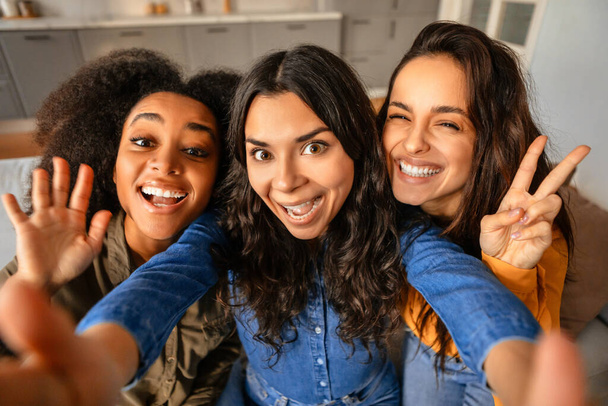 Três jovens multiculturais alegres tomando selfie brincalhão, acenando e mostrando sinais de paz, enquanto posam juntas, fazendo conteúdo de mídia social em casa. Beleza diversa e amizade - Foto, Imagem