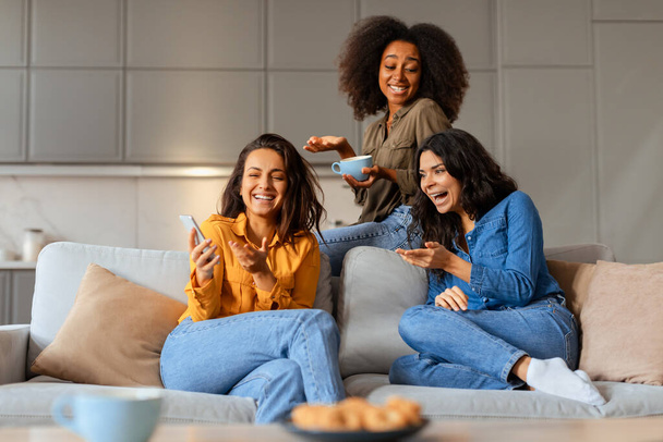 Tři veselé multiiraciální kamarádky sdílejí smích v obývacím pokoji, jedna drží mobilní telefon, vychutnává si kávu a relaxační víkend spolu, sedí na pohovce v interiéru - Fotografie, Obrázek