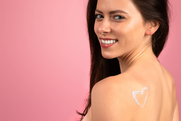 Jovem europeia feliz radiante com uma loção em forma de coração, aplicação de creme no ombro, sorrindo sobre o ombro com um olhar brincalhão, em um fundo rosa, close-up - Foto, Imagem