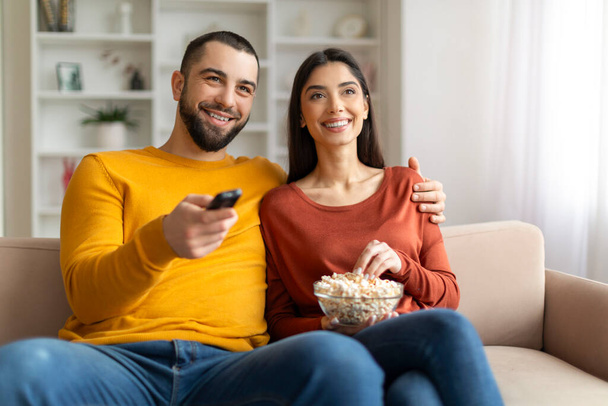 Rustig aan. Gelukkig jong getrouwd paar kijken tv in woonkamer, glimlachende Millennial echtgenoten eten samen popcorn terwijl ontspannen op comfortabele bank, Man Holding Remote Controller, close-up schot - Foto, afbeelding
