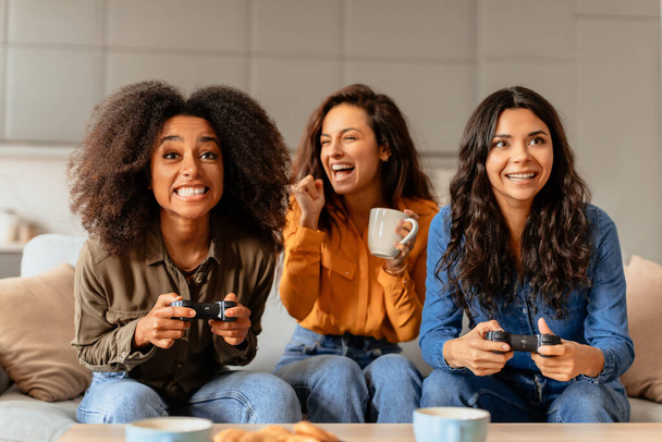 Multiculturali amiche giocano ai videogiochi nel loro accogliente salotto, godono di una sessione di gioco piena di divertimento, ridono impegnati in una competizione giocosa, seduti sul divano a casa al coperto - Foto, immagini