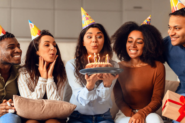 Весела група мультикультурних друзів, що збираються у вітальні, щоб відсвяткувати день народження, леді дме свічки на торт, роблячи її бажання на день народження, вдягаючи святкові капелюхи вдома. Святкування та дружба - Фото, зображення