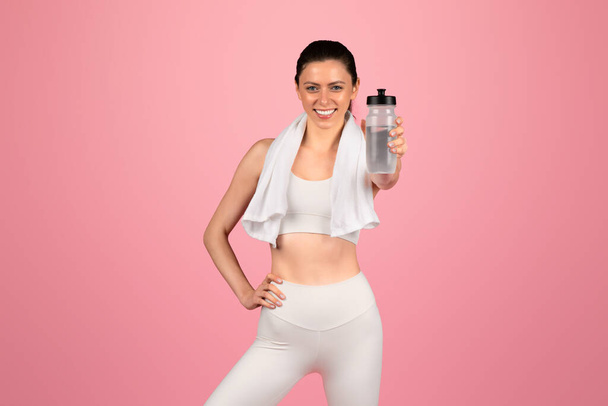 Λαμπερό χαμόγελο καυκάσιος νεαρή γυναίκα σε ένα λευκό αθλητικό σουτιέν και κολάν κρατώντας ένα μπουκάλι νερό με μια πετσέτα πάνω από τους ώμους της σε ροζ φόντο, προτείνουμε διατροφή, ξεκούραση - Φωτογραφία, εικόνα