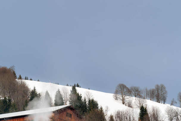 Eine ruhige Winterlandschaft mit einem schneebedeckten Hügel mit Bäumen und einer Hütte, die Rauch abgibt, unter einem klaren blauen Himmel. Ideal für Konzepte der Ruhe, Winter-Retreats und natürliche Schönheit - Foto, Bild