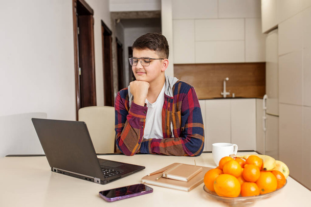 Enfocado adolescente con gafas está estudiando intensamente en su ordenador portátil, rodeado de materiales de estudio y fruta fresca en la mesa. General Z. - Foto, imagen