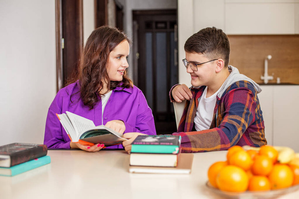 Δύο εστιασμένοι έφηβοι που ασχολούνται με την ανάγνωση και συζήτηση ακαδημαϊκού υλικού μαζί, με ένα σωρό βιβλία στο τραπέζι στο σπίτι περιβάλλον. - Φωτογραφία, εικόνα