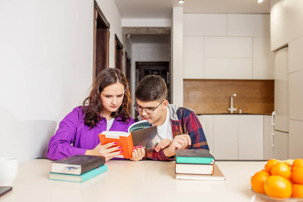 Δύο έφηβοι μαθητές εξετάζουν από κοντά το βιβλίο μαζί, που ασχολούνται με τη συλλογική μελέτη στο περιβάλλον στο σπίτι με στοίβα από βιβλία στο τραπέζι. - Φωτογραφία, εικόνα